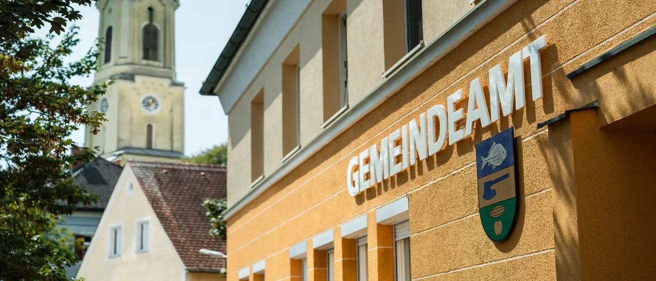 Ein gelbes Gebäude mit dem Wort Gemeindeam in Weiß auf der Vorderseite. Ein Kirchturm ist im Hintergrund sichtbar.