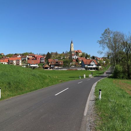 Eine Straße schlängelt sich durch ein Dorf mit rot gedeckten Häusern und einem Kirchturm unter einem klaren blauen Himmel. Grünes Gras und Bäume säumen die Straße.