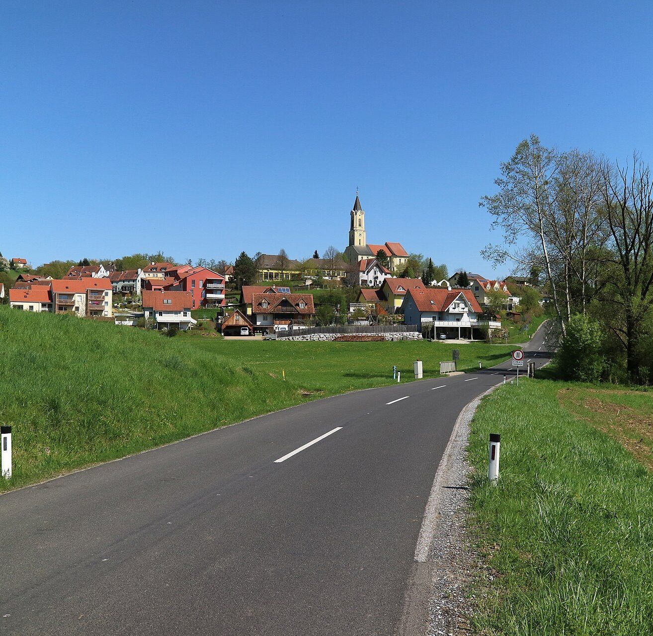 Eine Straße schlängelt sich durch ein Dorf mit rot gedeckten Häusern und einem Kirchturm unter einem klaren blauen Himmel. Grünes Gras und Bäume säumen die Straße.