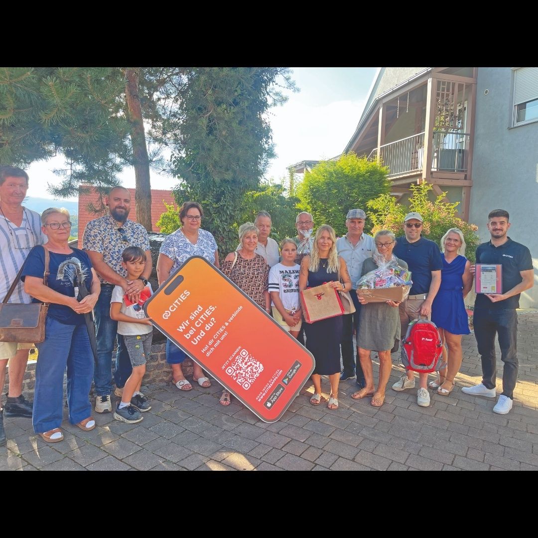 Eine Gruppe von Menschen, darunter Erwachsene und Kinder, steht vor einem Gebäude, hält Geschenke und lächelt für ein Foto. Ein großes orangefarbenes Plakat mit dem Text 'Wir sind die Städte. Und du?' ist prominent zu sehen.