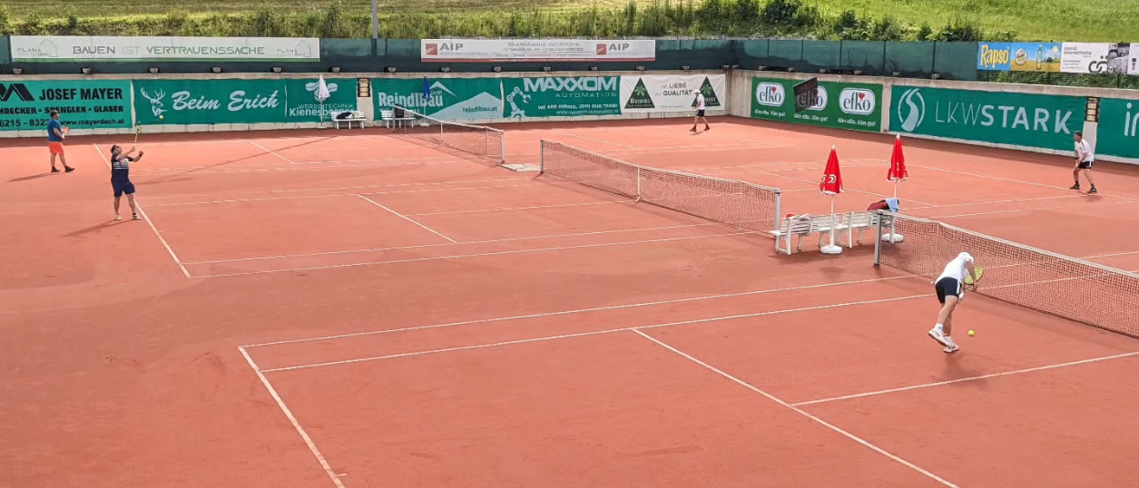 Mehrere Tennisspieler spielen auf einem roten Sandplatz mit grünen Wänden und Bannern. Ein Spieler beugt sich in der Nähe des Netzes.