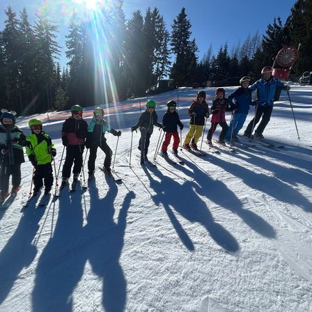 Eine Gruppe junger Skifahrer in Winterkleidung und Helmen posiert auf einem schneebedeckten Hang mit immergrünen Bäumen im Hintergrund.