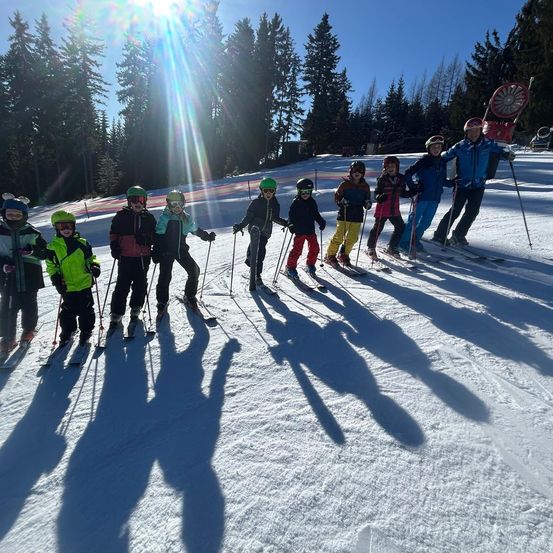 Eine Gruppe junger Skifahrer in Winterkleidung und Helmen posiert auf einem schneebedeckten Hang mit immergrünen Bäumen im Hintergrund.