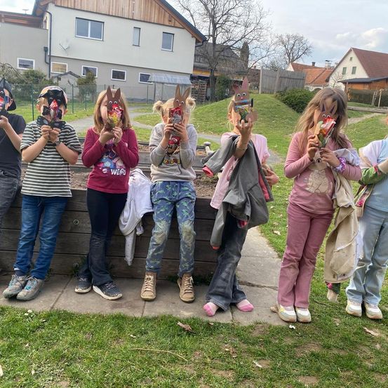 Eine Gruppe von Kindern, alle mit Hasenmasken, stehen auf einem Betonweg, halten einige Snacks und posieren für ein Foto. Sie scheinen sich in einem Park mit grünem Rasen und Häusern im Hintergrund zu befinden.