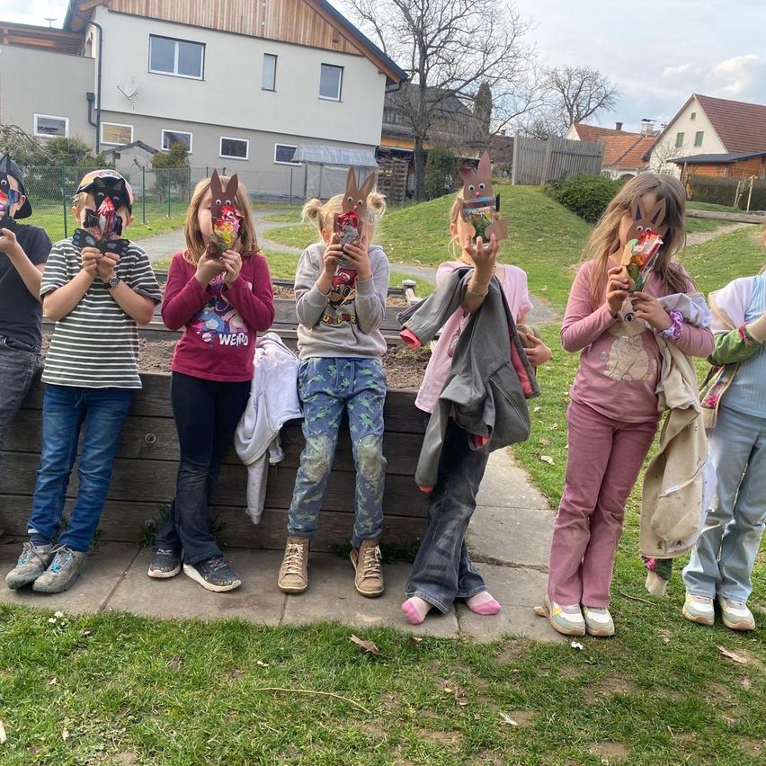 Eine Gruppe von Kindern, alle mit Hasenmasken, stehen auf einem Betonweg, halten einige Snacks und posieren für ein Foto. Sie scheinen sich in einem Park mit grünem Rasen und Häusern im Hintergrund zu befinden.