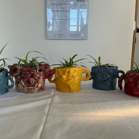 Fünf einzigartige Keramikbecher mit Pflanzen auf einer weißen Tischdecke. Ein weißes Schild ist an der Wand dahinter.