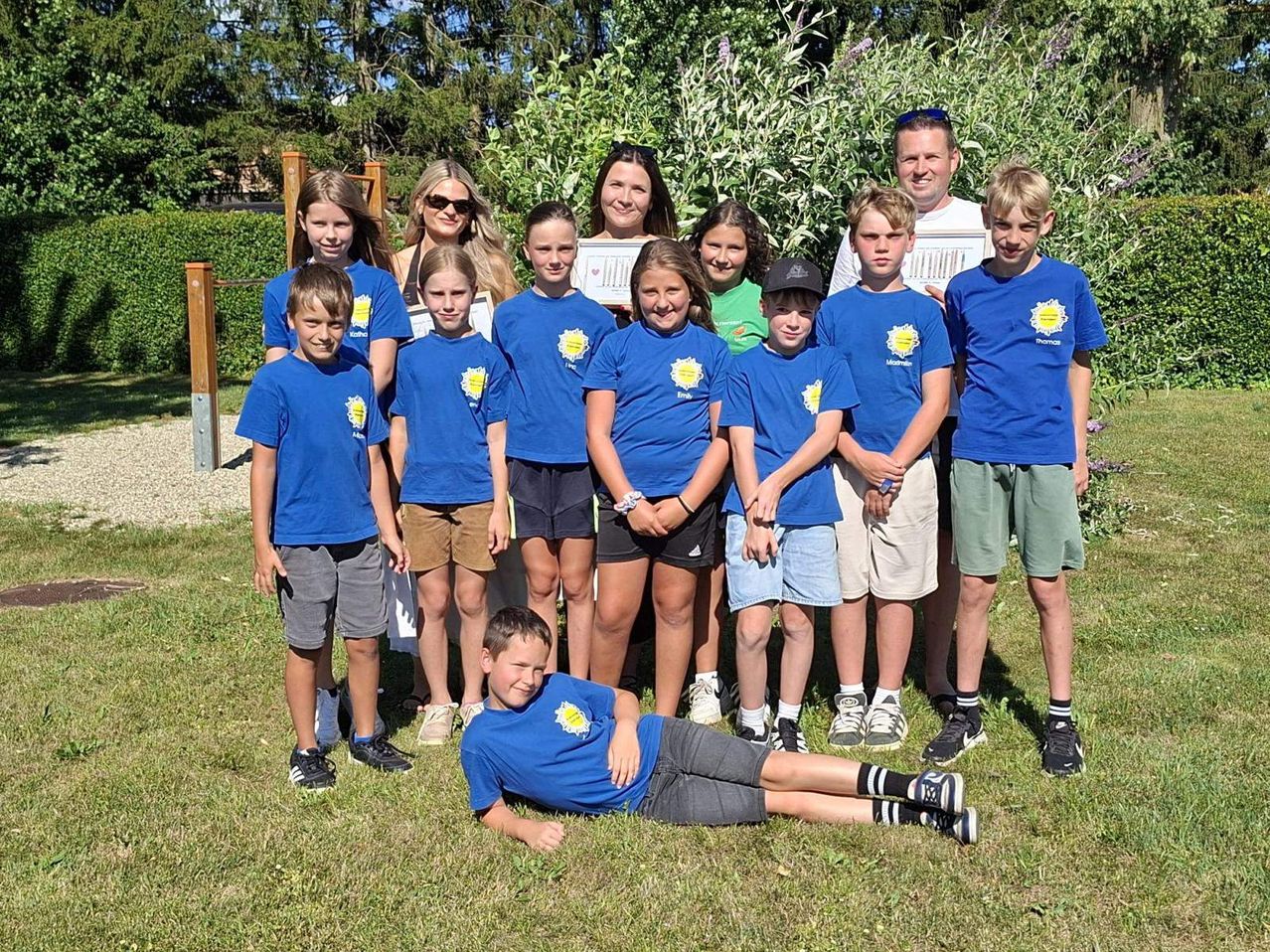 Eine Gruppe von Kindern in blauen T-Shirts und einige Erwachsene stehen in einem Garten. Sie haben Zertifikate und scheinen zu feiern.