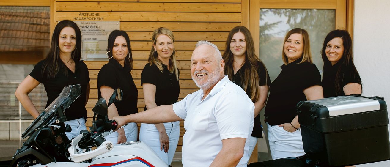 Ein Mann und sieben Frauen stehen zusammen vor einem Gebäude mit einem Schild für Dr. Med. Uny Hausapotheke. Der Mann sitzt auf einem BMW Motorrad und lächelt. Die Frauen tragen schwarze Shirts und weiße Hosen.