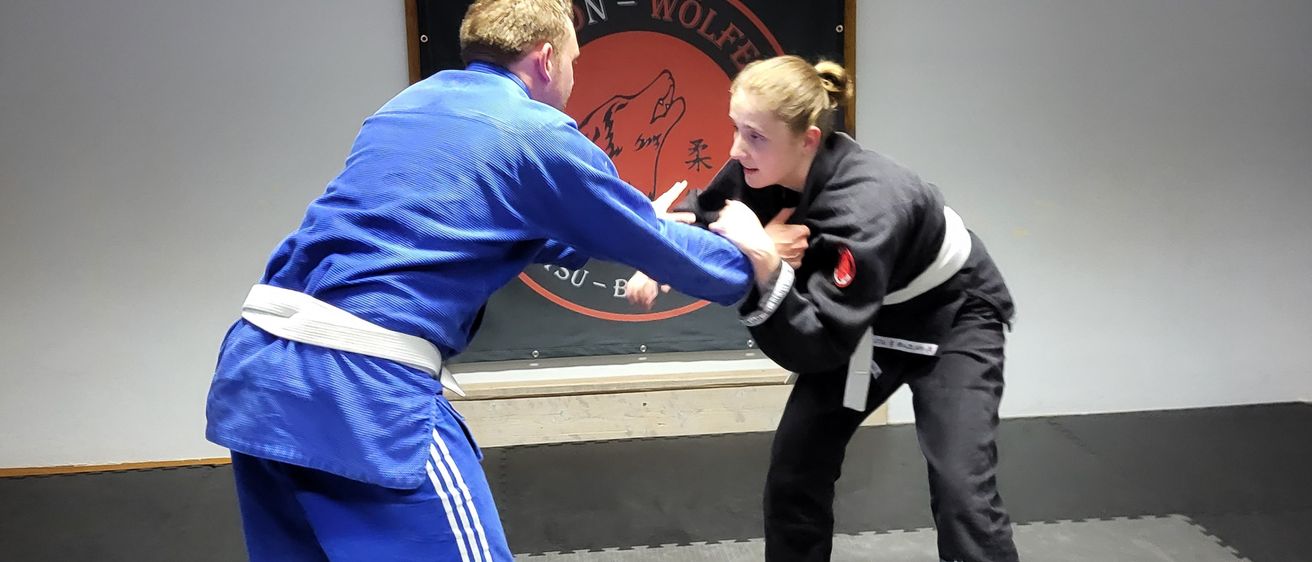 Two individuals are practicing martial arts in a room with a white wall and a black banner with text and a logo. The man is in a blue gi and the woman is in a black gi.