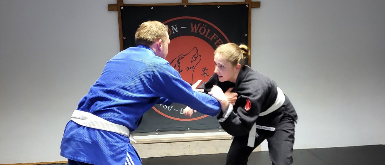 Zwei Personen üben Kampfsport in einem Raum mit weißer Wand und schwarzem Banner mit Text und Logo. Der Mann trägt ein blaues Gi und die Frau ein schwarzes Gi.