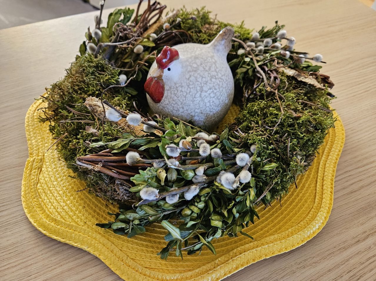 Eine Keramik-Henne sitzt in einem Nest aus grünem Moos und Zweigen auf einem gelben Untersetzer.