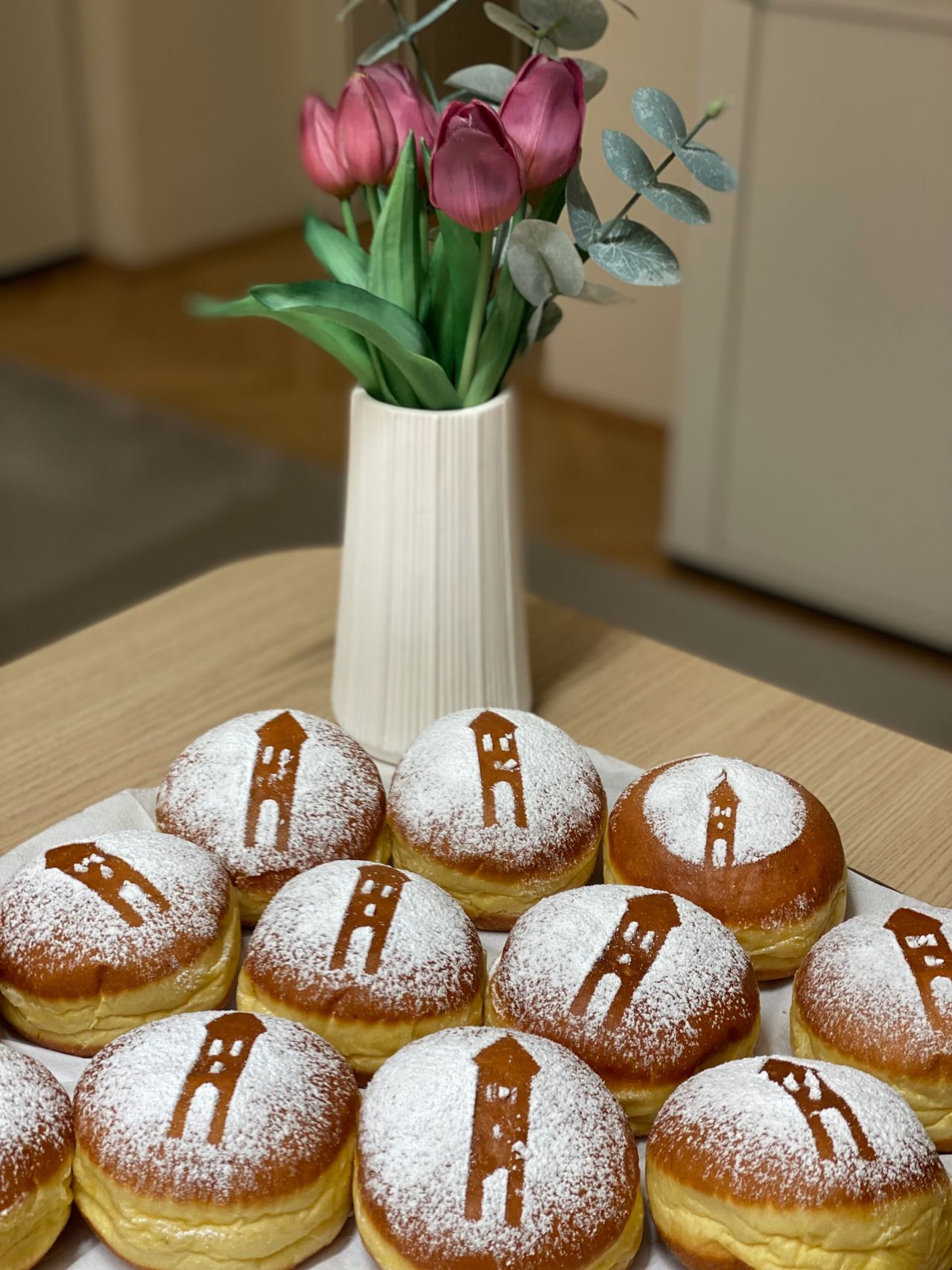 Eine Platte mit puderzuckerüberzogenen Donuts mit einer Zeichnung eines Hauses darauf, begleitet von einer weißen Vase mit rosa Tulpen und Eukalyptusblättern.