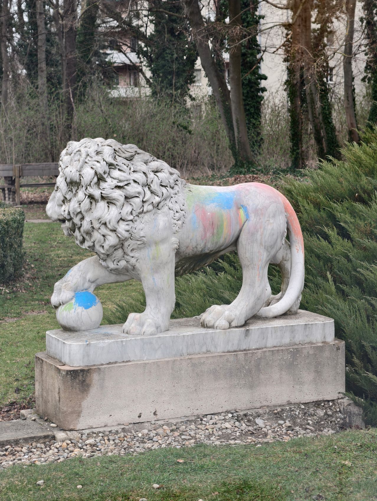 Eine Löwenstatue mit mehrfarbiger Bemalung auf dem Körper steht auf einem Betonsockel in einem Park. Der Löwe hält einen Ball in seinen Pfoten.