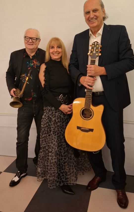 Drei Menschen stehen zusammen, zwei Männer und eine Frau, lächeln und posieren für ein Foto. Ein Mann hält eine Trompete, der andere eine Gitarre, und die Frau trägt einen Leopardenprintrock.