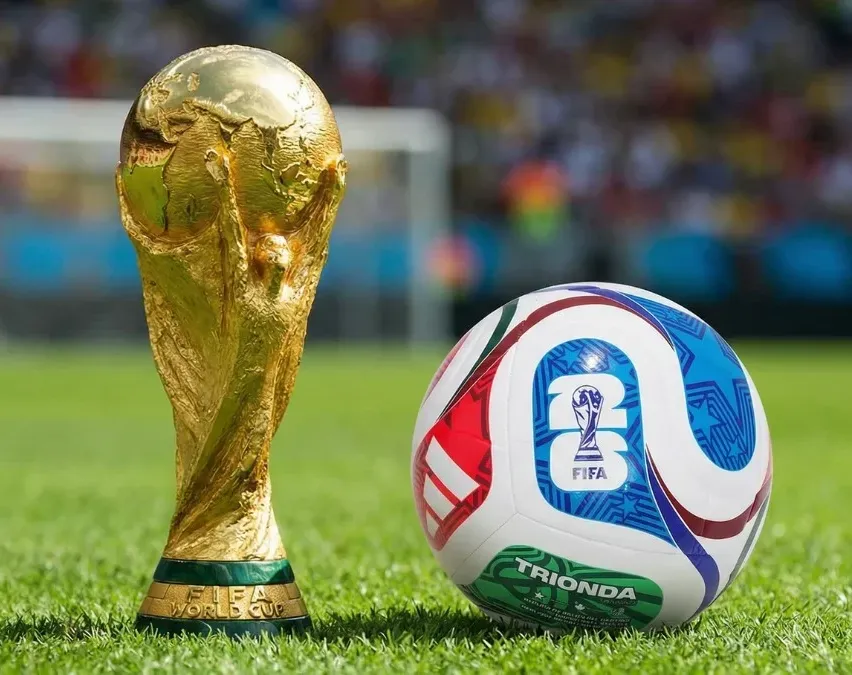 Ein goldener FIFA-Weltmeister-Pokal neben einem Fußball mit der Nummer 23 auf einem grünen Feld.
