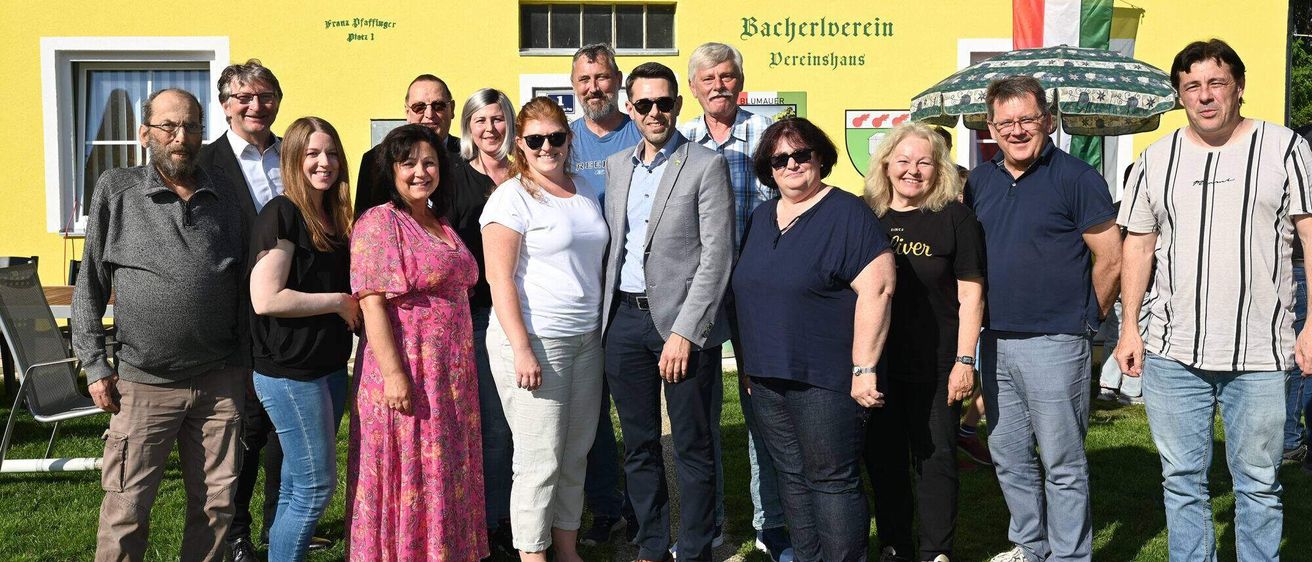 Eine Gruppe von Menschen steht vor einem gelben Gebäude mit dem Text 'Bacherlverein'. Sie tragen Freizeitkleidung und lächeln. Hinter ihnen steht ein grüner Regenschirm.
