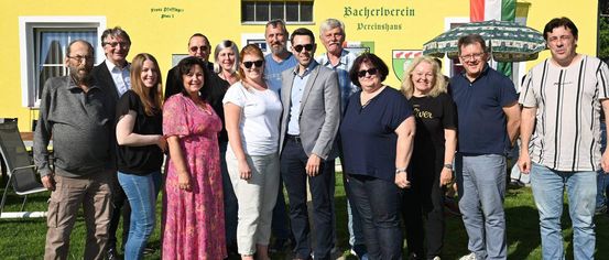 A group of people stands in front of a yellow building with the text 'Bacherlverein'. They are wearing casual clothes and smiling. Behind them is a green umbrella.