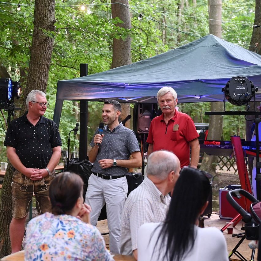 Eine Gruppe von Menschen steht unter einem Zelt im Wald, wobei ein Mann ein Mikrofon hält und spricht. Ein weiterer Mann steht neben ihm, und einige andere hören zu. In der Nähe ist Musikequipment aufgebaut.