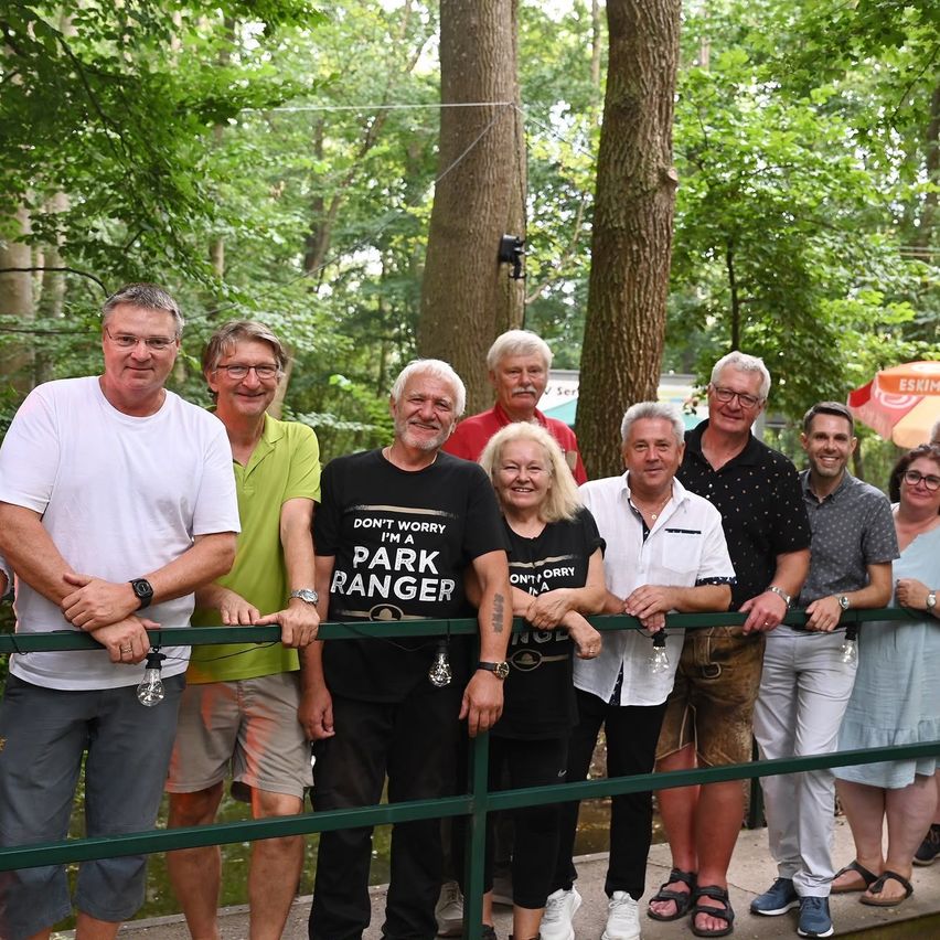 Eine Gruppe von Erwachsenen, darunter Männer und Frauen, stehen lächelnd für ein Foto in einem Wald. Einige tragen Brillen, und ein Mann hat ein 'Park Ranger'-Shirt an. Sie stehen hinter einem grünen Geländer.