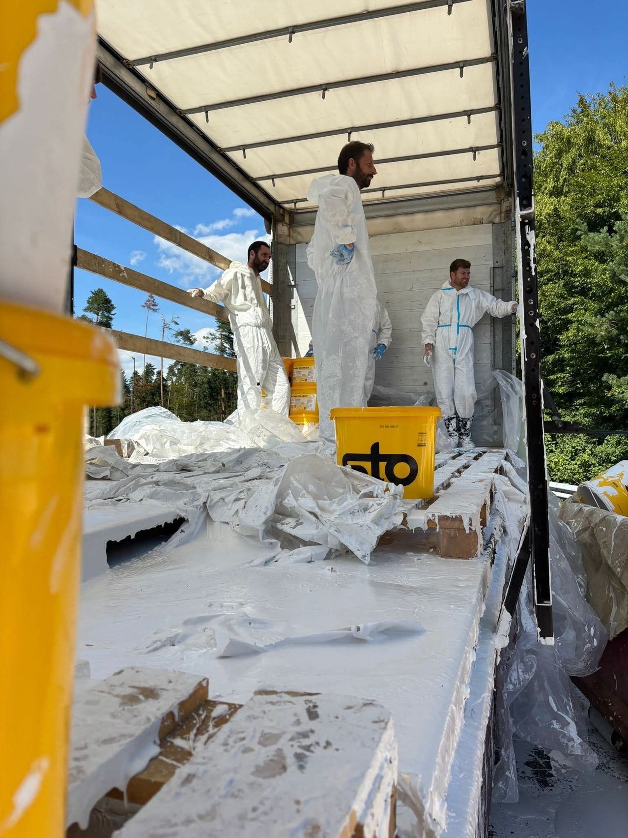 Drei Männer in weißen Schutzanzügen arbeiten in einem mit Plastiktüten und gelben Behältern gefüllten LKW, möglicherweise bei der Aufräumung eines Unfalls.