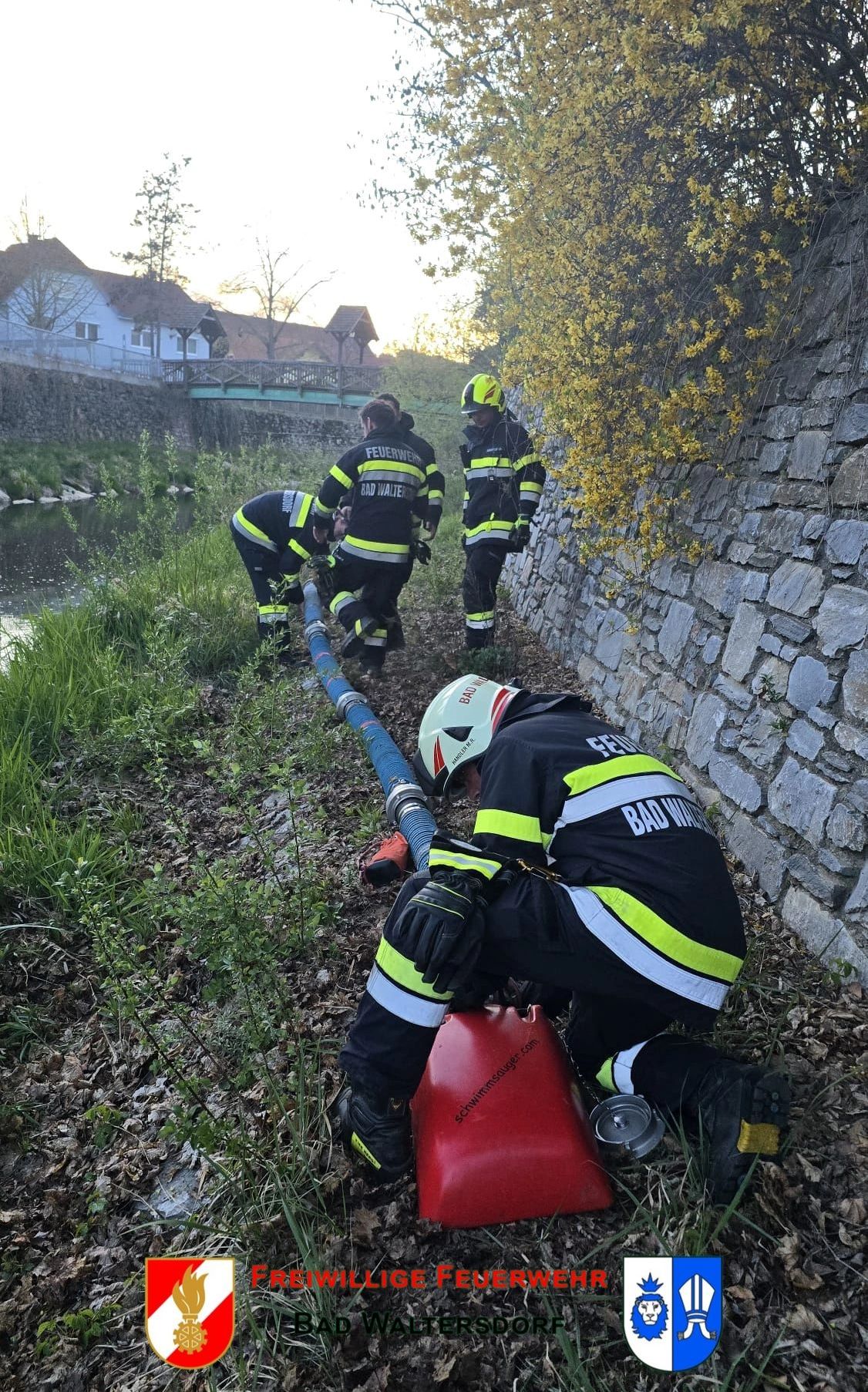 Firefighters in uniform are working near a river, one is crouching down with a red tank, while others stand with a hose.