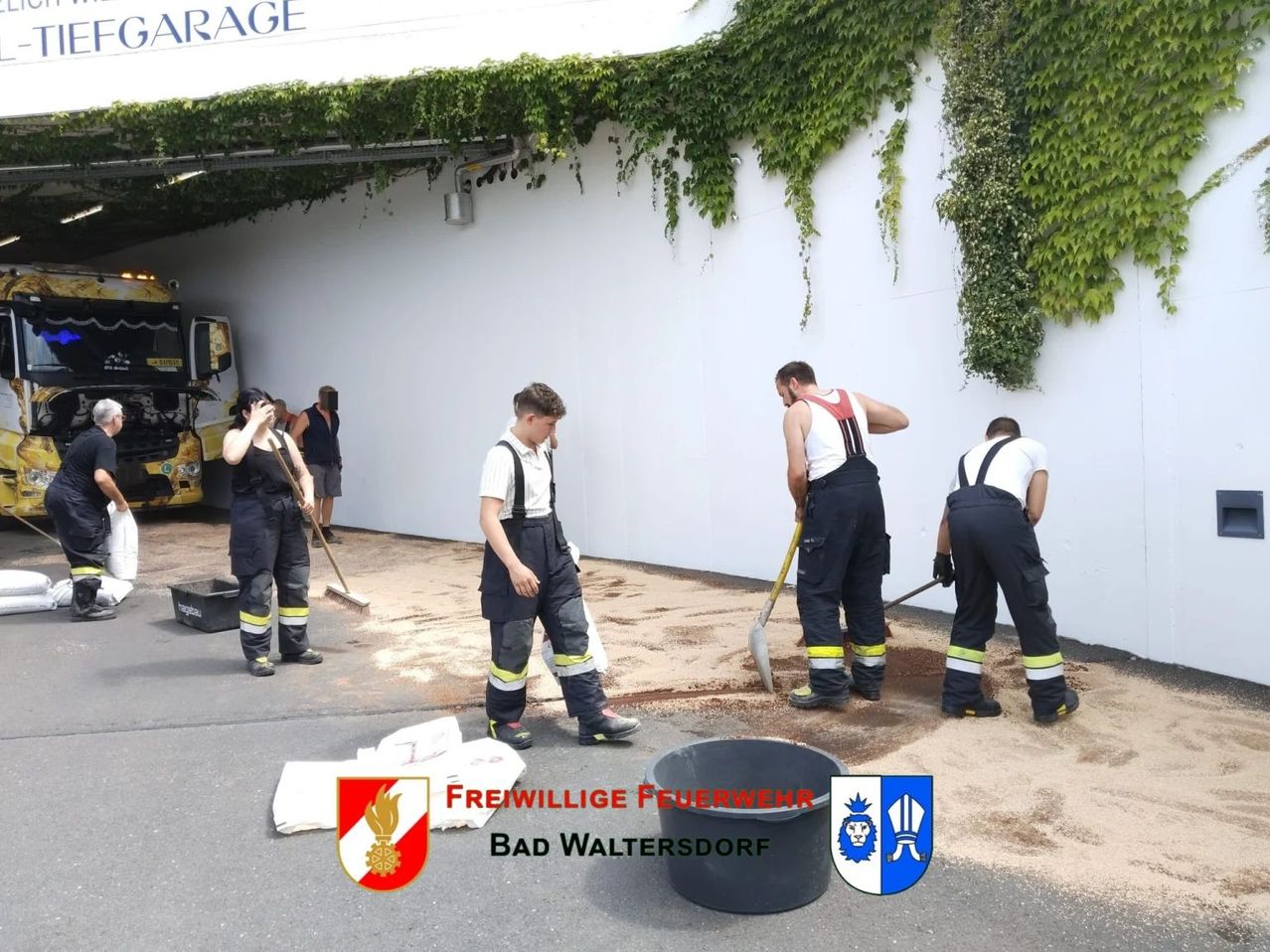 Freiwillige Feuerwehrleute in Bad Waltersdorf säubern einen Schmutzbereich, einer hält einen Besen und andere benutzen Schaufeln. Die Szene spielt vor einer mit Efeu bewachsenen weißen Wand.