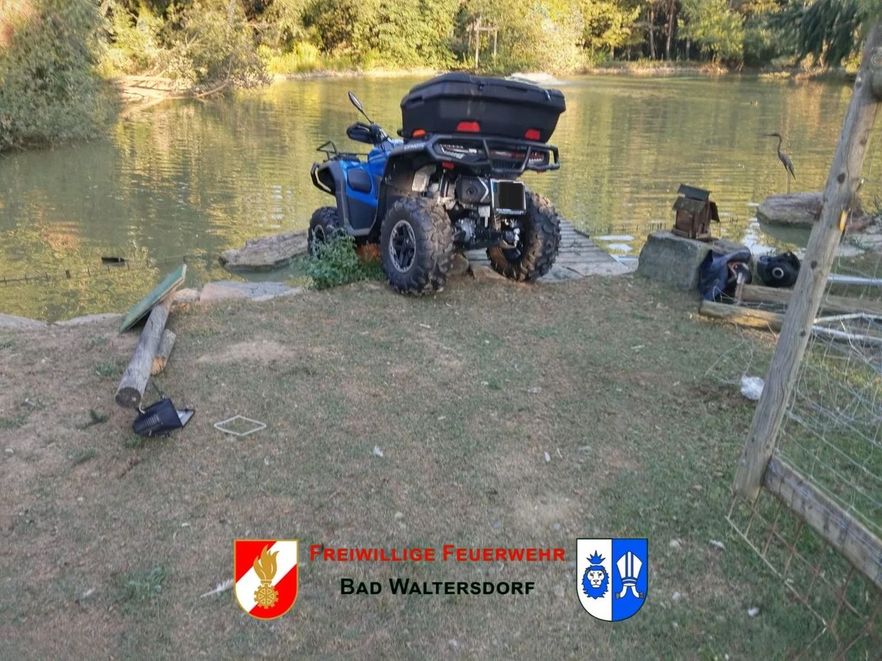 A blue ATV is parked on the grass near a lake with a storage box on its back. Nearby, there are some rocks, a wooden log, and a small wooden box. The image also includes logos for Freiwillige Feuerwehr Bad Waltersdorf.