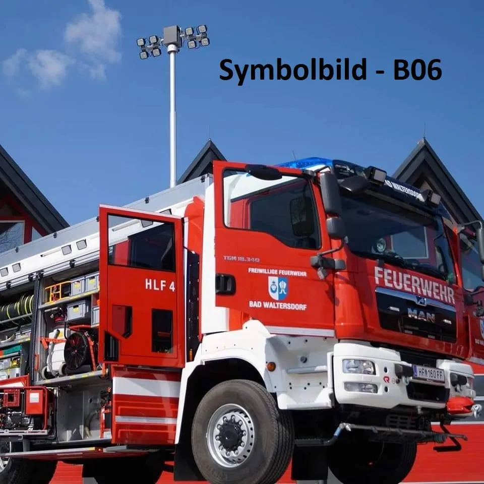 Ein roter und weißer Feuerwehrwagen mit der Aufschrift HLF 4, geparkt vor einem roten Gebäude mit klarem Himmel und einer Straßenlaterne im Hintergrund.