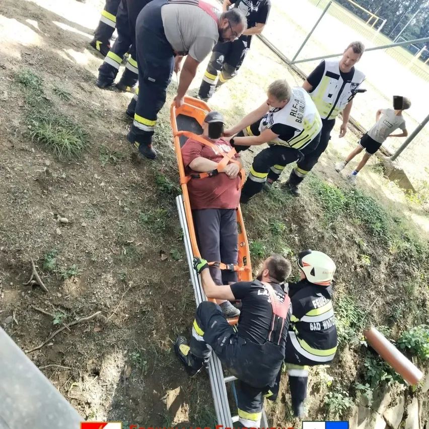 Feuerwehrleute senken vorsichtig eine Person auf einer Trage einen steilen Hang hinunter. Sie tragen Helme und Uniformen.