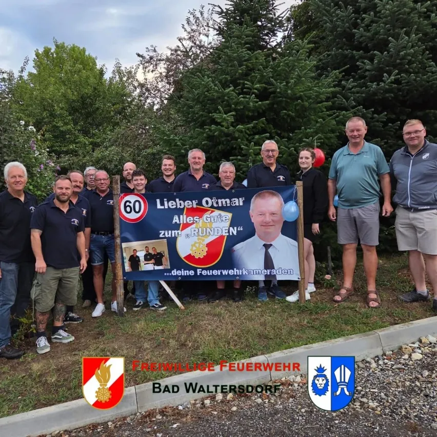 Eine Gruppe von Menschen, einschließlich Feuerwehrleuten, steht vor einem Banner, das einen 60. Geburtstag feiert. Das Banner zeigt ein Foto eines Mannes und die Worte 'Lieber Otmar Alles Gute zum RUNDEN'. Die Gruppe steht auf Gras mit Bäumen im Hintergrund.