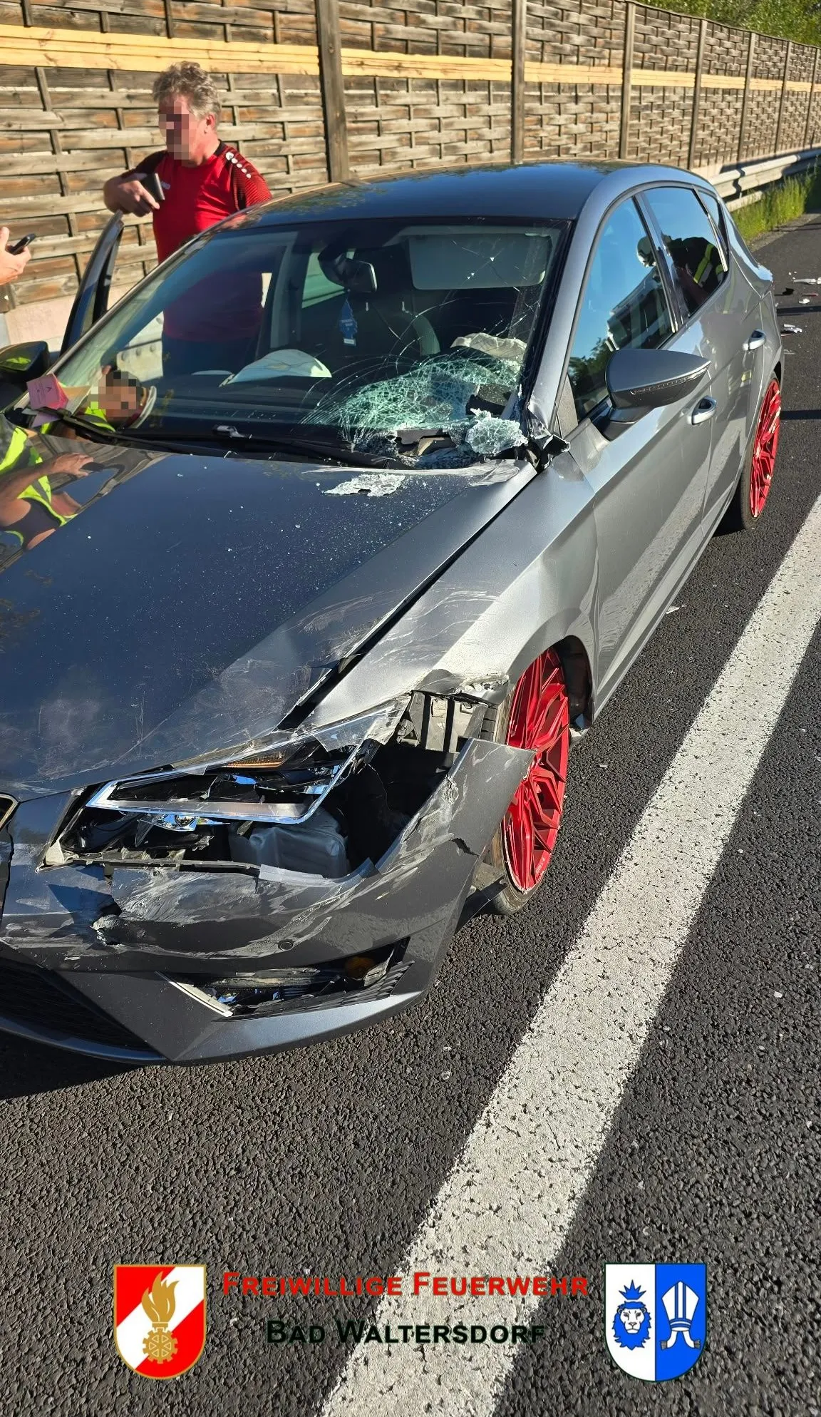 Ein beschädigtes graues Auto mit zerbrochener Windschutzscheibe und roten Felgen steht am Straßenrand.