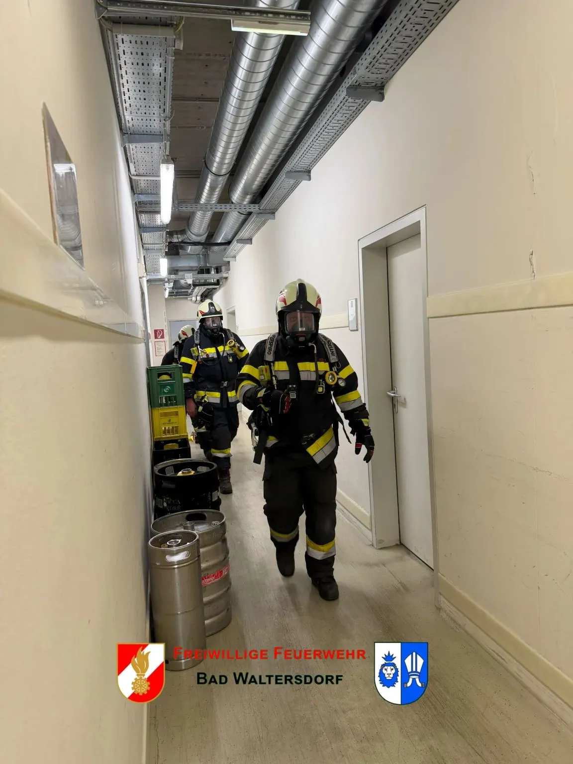 Drei Feuerwehrleute in voller Ausrüstung gehen einen Flur entlang mit Metallrohren an der Decke und Bierfässern an der Seite.