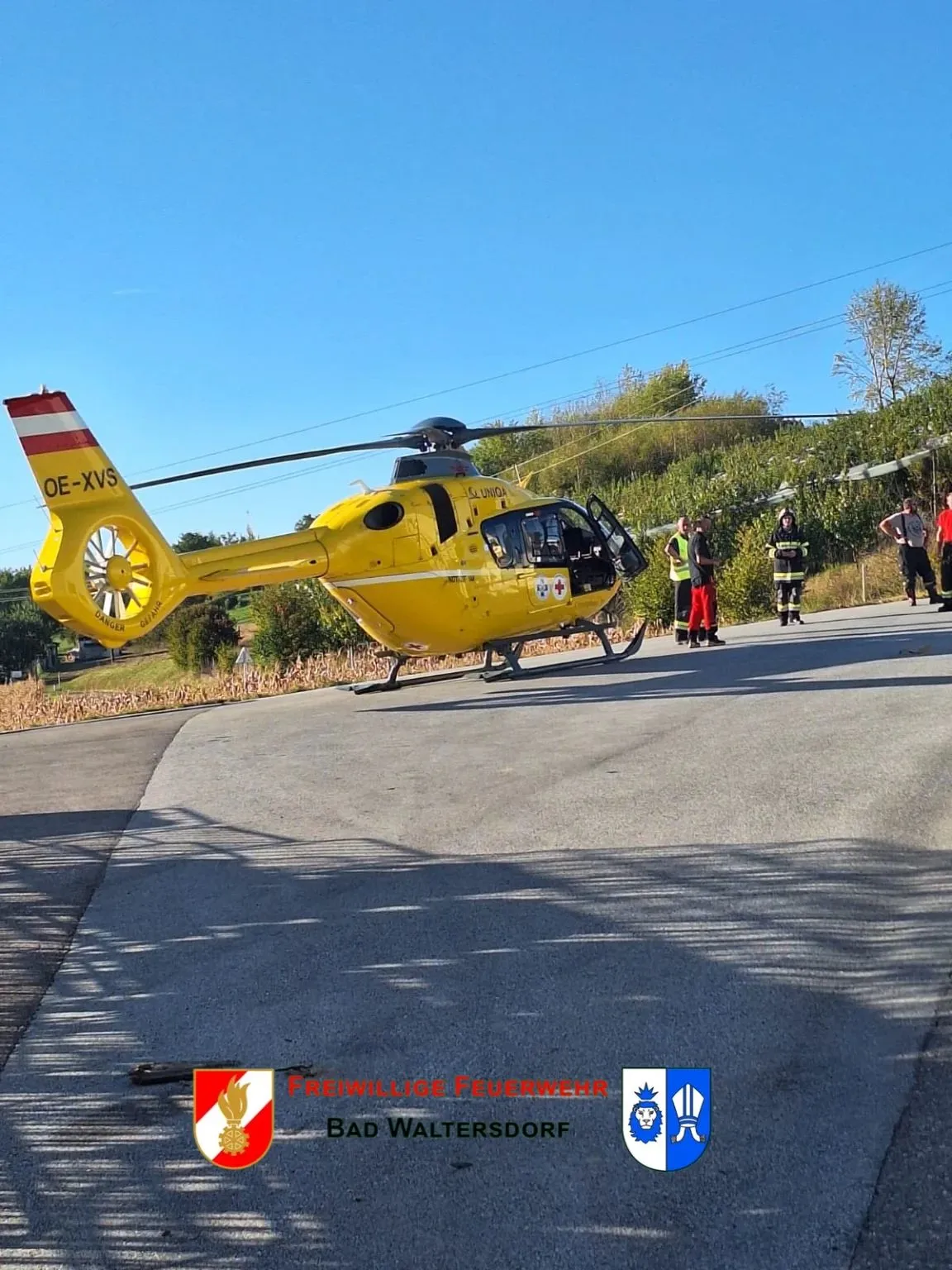 Ein gelber Hubschrauber mit der Registrierung OE-XVS steht auf der Straße, mehrere Personen in Sicherheitsausrüstung stehen in der Nähe.