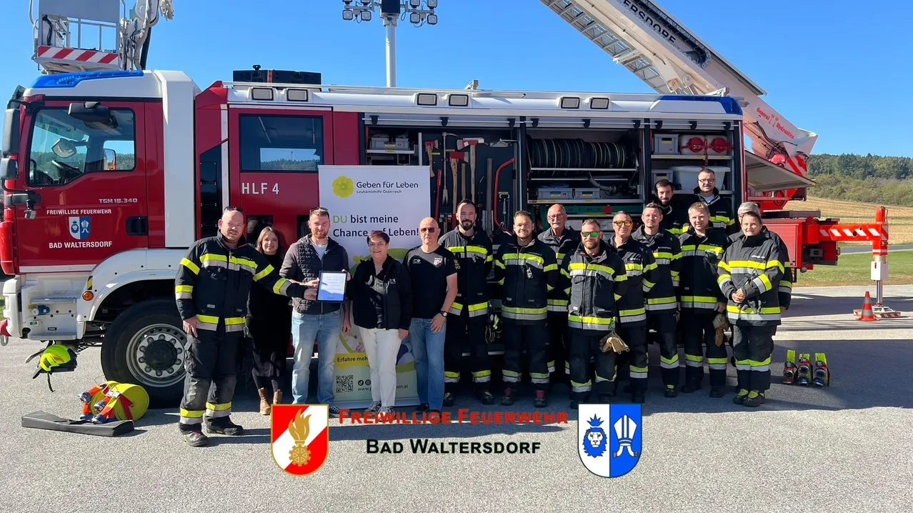 Eine Gruppe von Feuerwehrleuten in Uniform steht vor einem Feuerwehrauto mit der Aufschrift 'HLF 4'. Sie posieren für ein Foto mit einer Frau, die ein Zertifikat in der Hand hält.