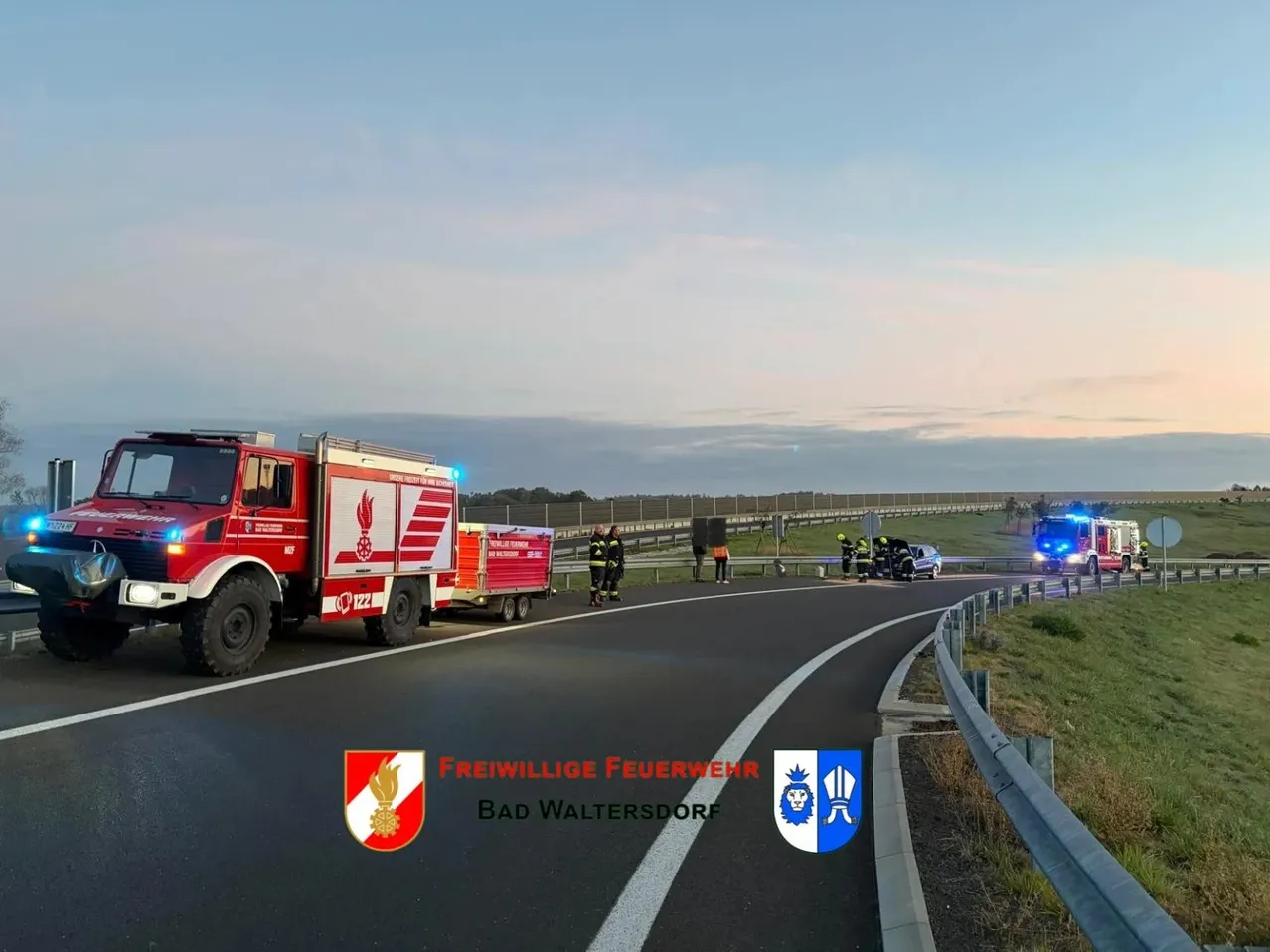 Ein Feuerwehrauto ist am Straßenrand geparkt, mit blinkenden Notleuchten. Andere Rettungsfahrzeuge sind in der Nähe, und Menschen stehen um ein Auto herum, das in einen Unfall verwickelt war.