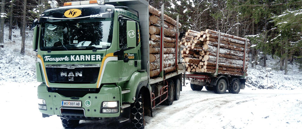 Ein grüner Lastwagen mit dem Namen Karner ist mit Baumstämmen beladen und fährt auf einer verschneiten Straße.