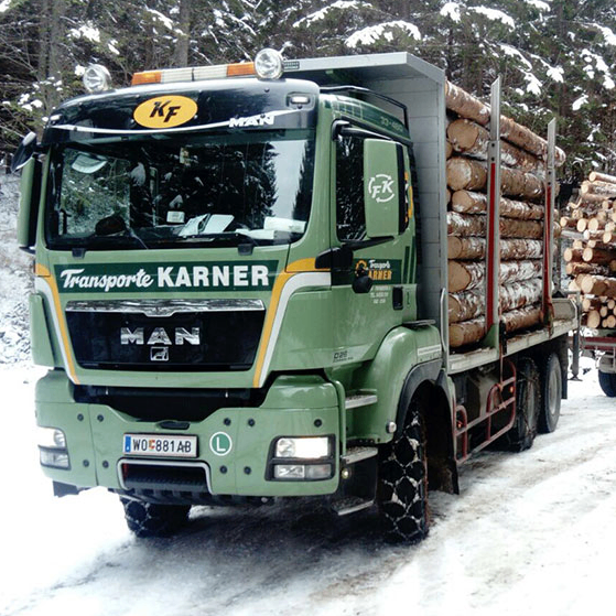 Ein grüner Lastwagen mit Baumstämmen fährt durch einen verschneiten Wald. Er ist mit Karner Transport beschriftet.