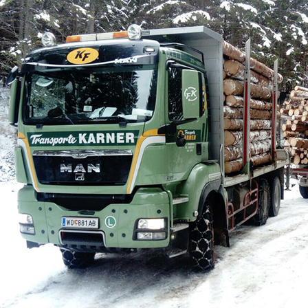 Ein grüner Lastwagen mit Baumstämmen fährt durch einen verschneiten Wald. Er ist mit Karner Transport beschriftet.