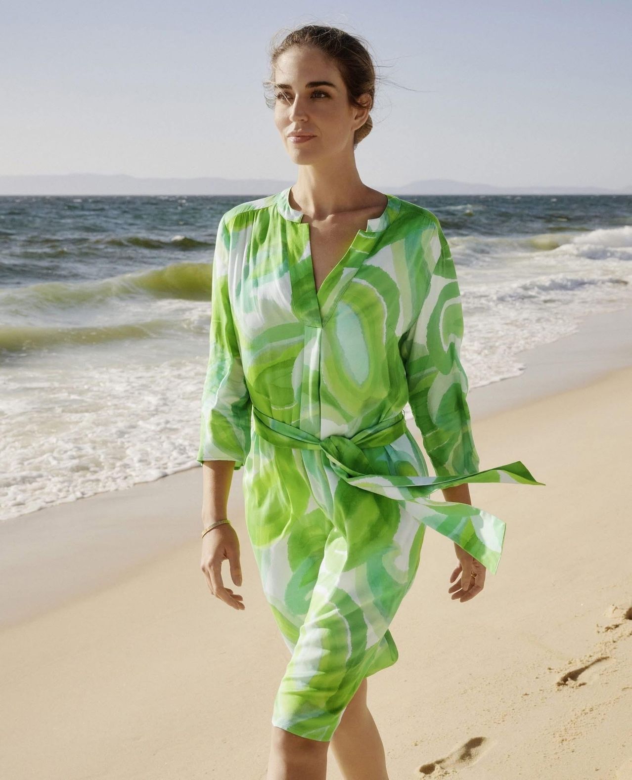 Eine Frau in einem grünen Kleid geht am Strand entlang, mit Wellen im Hintergrund.