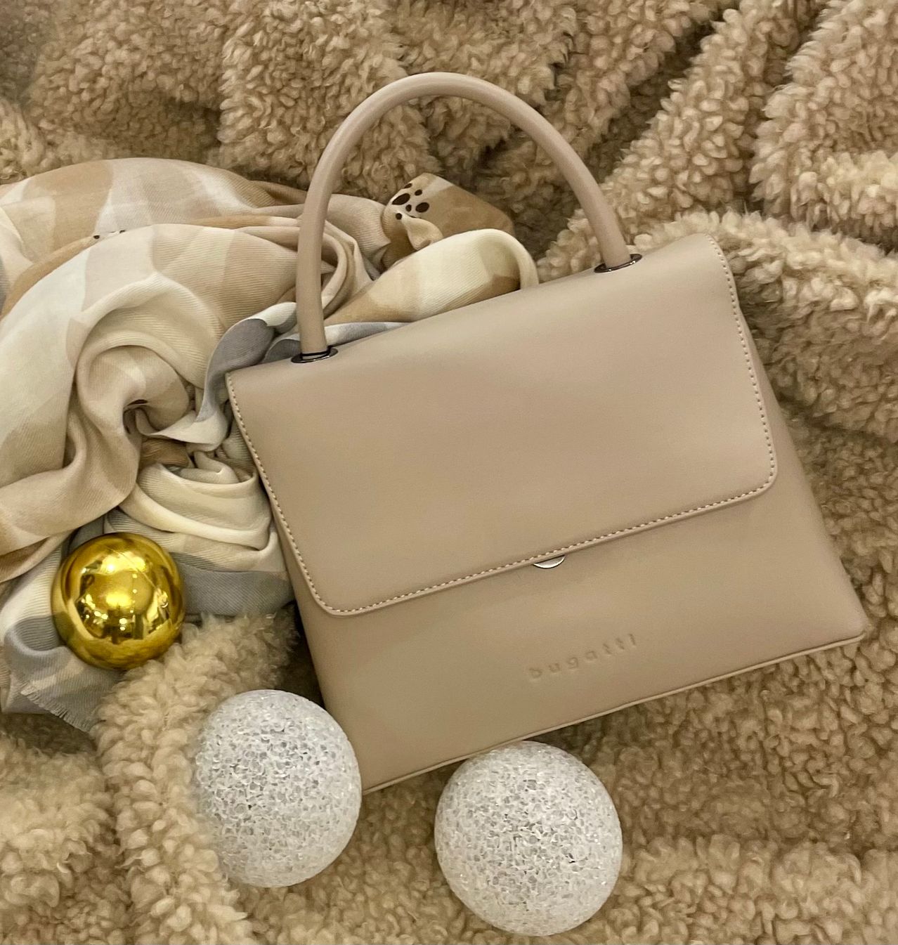 A beige Bugatti handbag rests on a fluffy carpet with a scarf and two glass balls, one golden and one white.