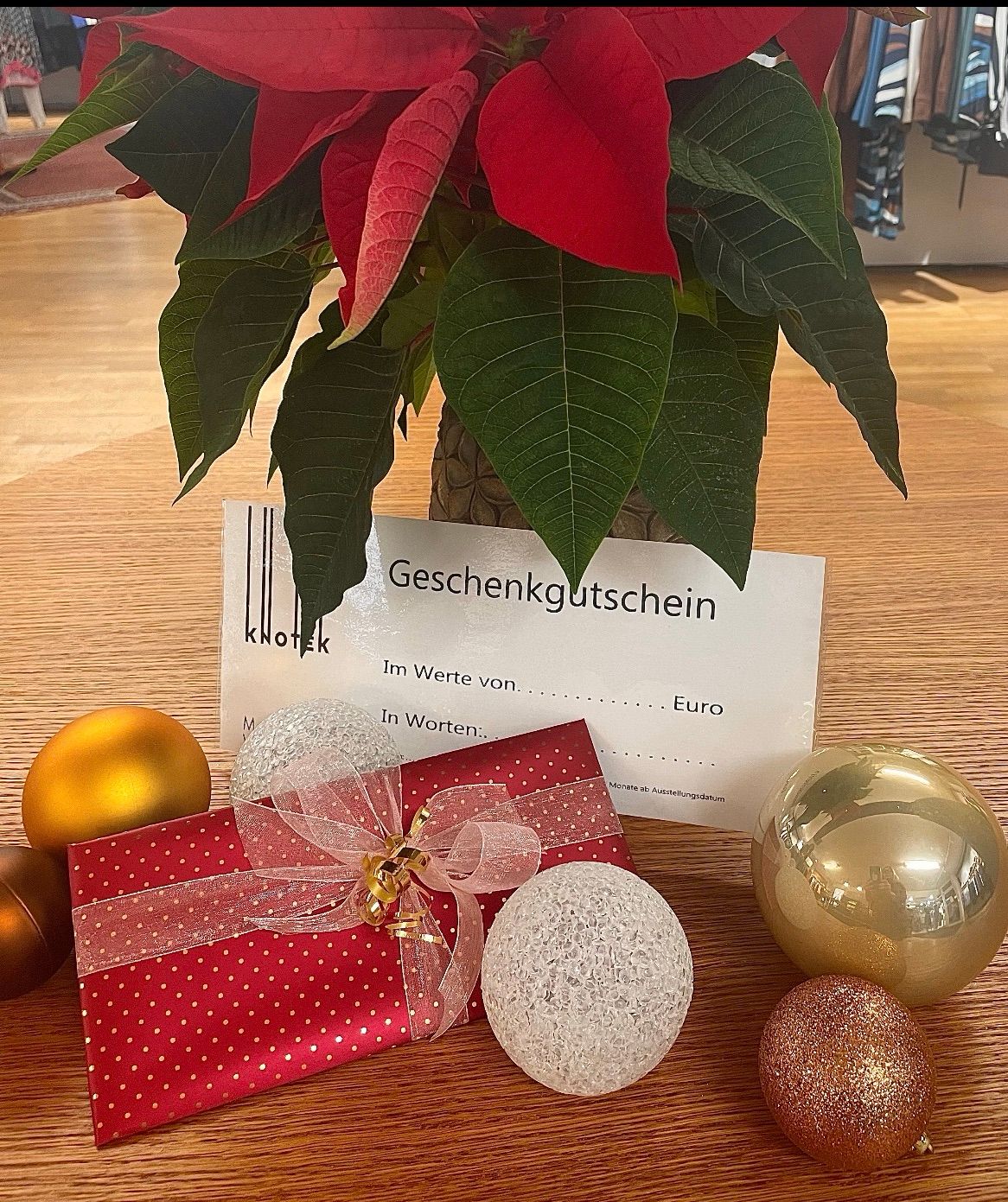 Ein rotes Geschenkbox mit einem Band liegt auf einem Holztisch mit einem Geschenkgutschein und dekorativen Kugeln. Hinter dem Geschenkbox steht eine rote Weihnachtsstern-Pflanze in einem braunen Topf.