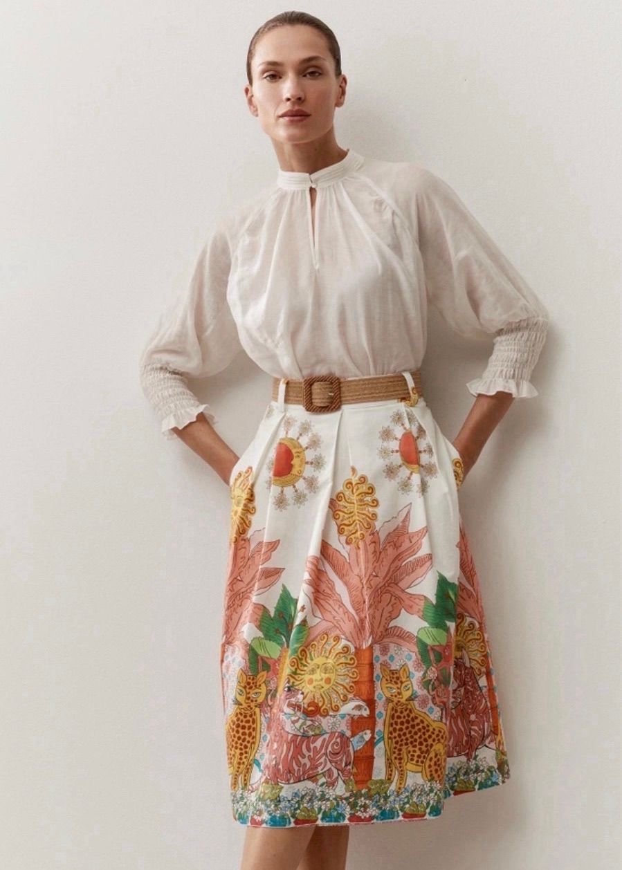 Eine Frau steht und trägt ein weißes Oberteil mit Rüschen und einen Blumenrock. Sie hat einen braunen Gürtel an.