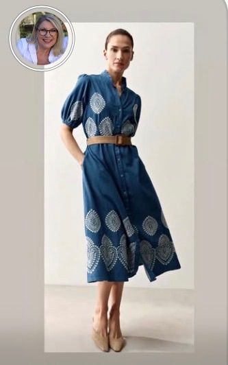 Eine Frau in einem blau bedruckten Kleid mit einem braunen Gürtel posiert drinnen vor einem weißen Hintergrund.