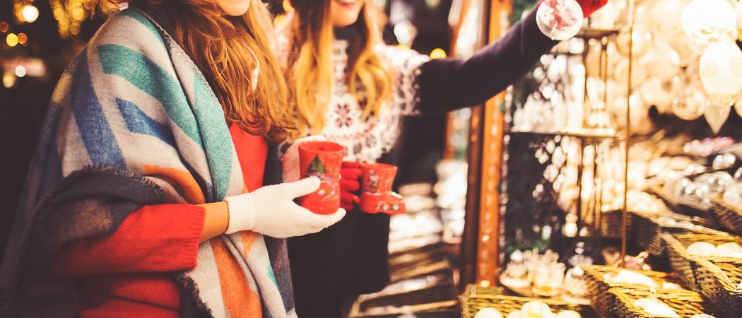 Zwei Frauen in Winterkleidung halten rote Tassen, möglicherweise auf einem Weihnachtsmarkt, mit einem verschwommenen Hintergrund aus Holzregalen und Lichtern.