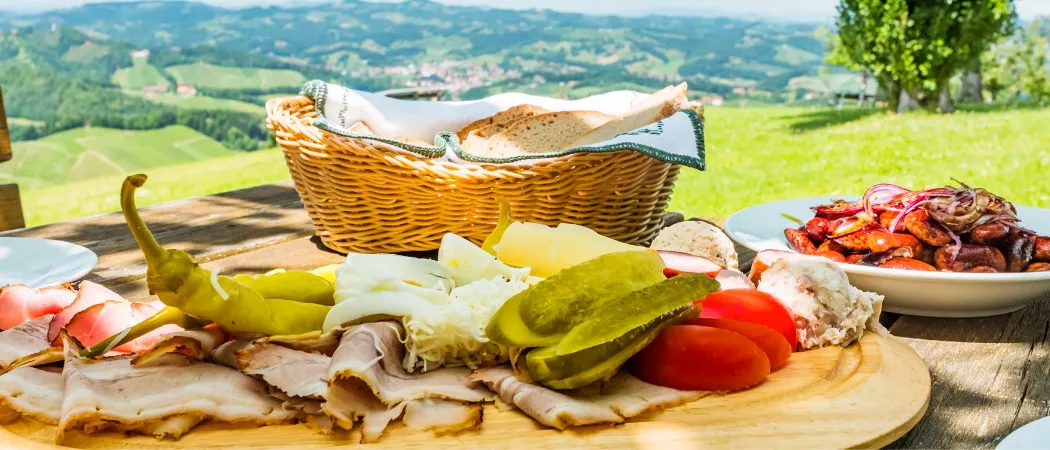 Ein Picknick-Setup im Freien mit einem Holzbrett voller Fleisch, Käse, Gurken und Tomaten, mit einem Korb Brot und einem Blick auf ein Tal im Hintergrund.