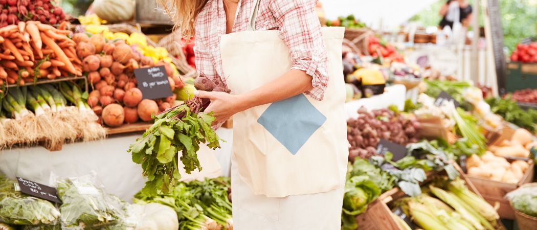 Eine Frau auf einem Markt hält eine Handvoll Rüben in einer Hand und eine Tasche in der anderen. Sie trägt eine weiße Schürze und ein kariertes Hemd.