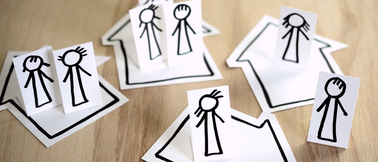 Mehrere ausgeschnittene Familienfiguren aus Streichholz auf einer Holzoberfläche, jede in einer Hausform, einige mit Gesichtern und einige ohne.