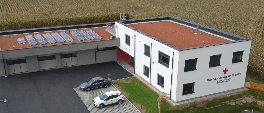 Luftaufnahme des Osterreichischen Roten Kreuz Steiermark Gebäudes mit rotem Dach, Solarpanelen und einem Parkplatz mit drei Autos.