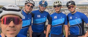 Fünf Männer in Radfahrer-Ausrüstung und Helmen stehen in einer Reihe. Sie lächeln und posieren für ein Foto.