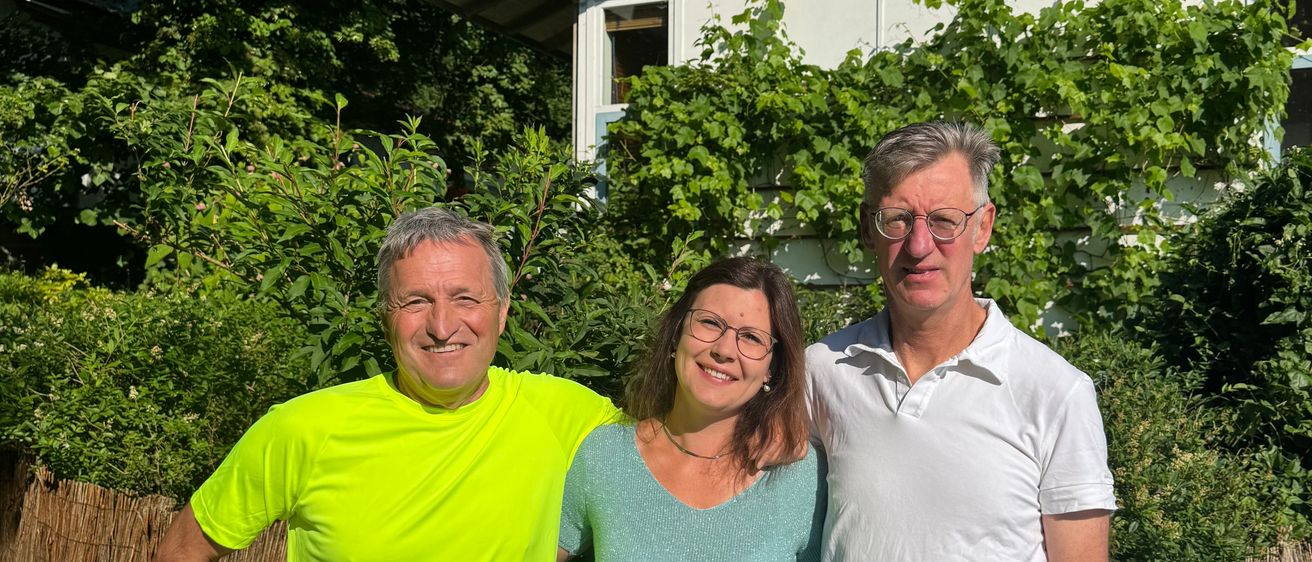 Drei Personen stehen vor einem Haus mit einem braunen Dach, umgeben von viel Grün. Ein Mann in einem neongelben Shirt, eine Frau in einem türkisen Top und ein weiterer Mann in einem weißen Polohemd.